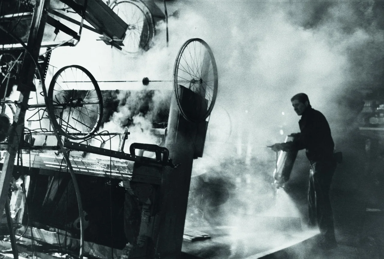 Smoke surrounds the wheels and machinery of Homage to New York, with a person standing at the right.