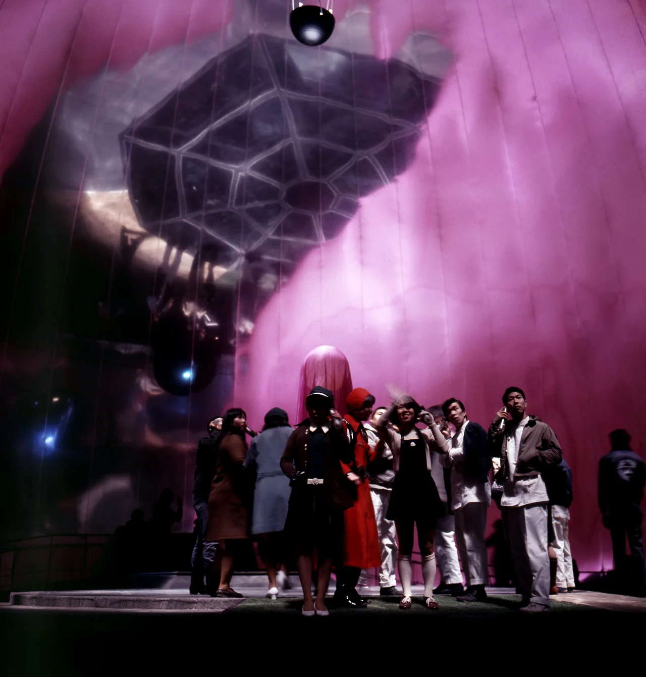 Visitors holding handsets stand beneath pink light and a suspended balloon in the Pepsi Pavilion mirror room.