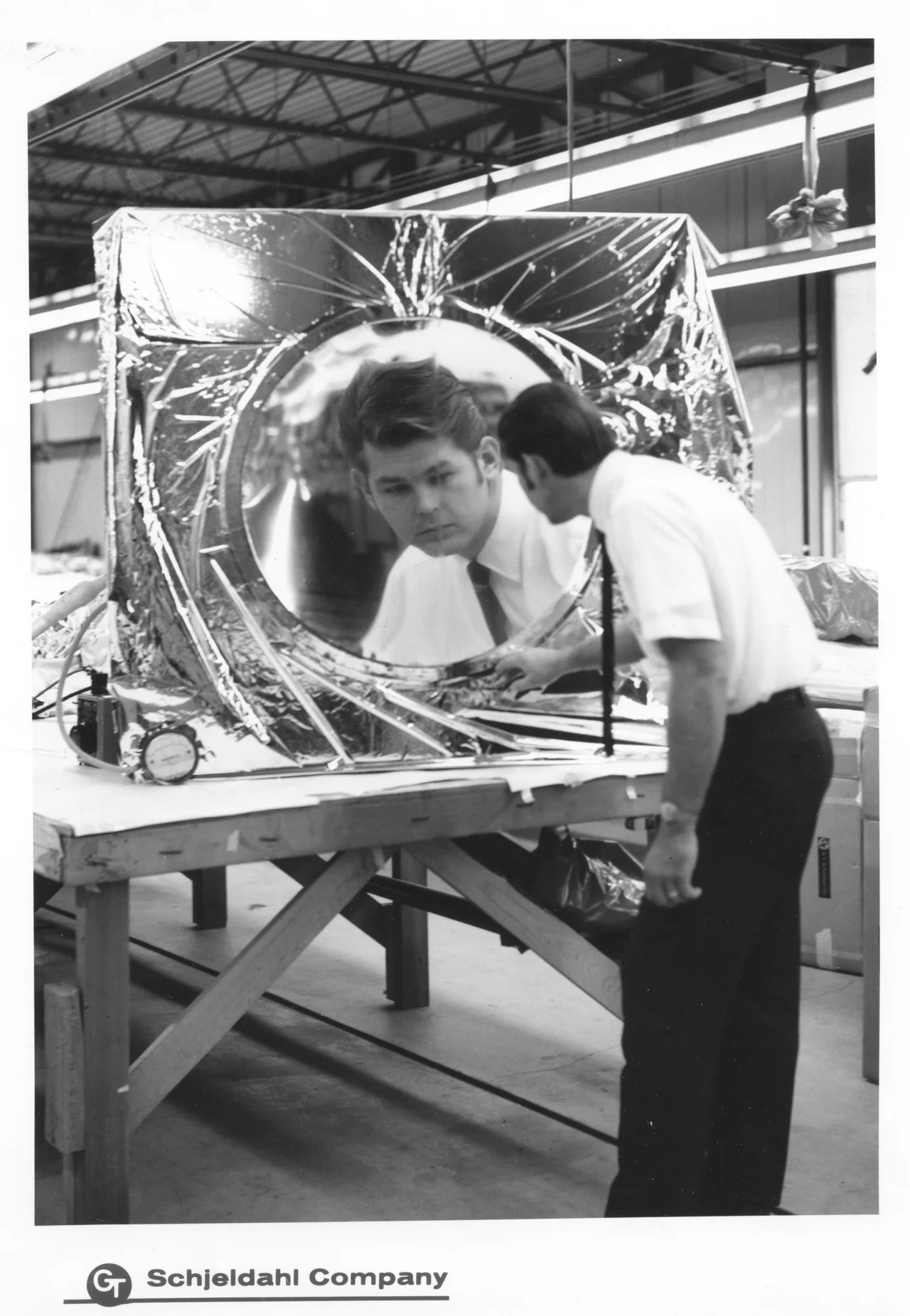 A man looks into a small curved mirror on a workshop table; his enlarged reflection fills its center.
