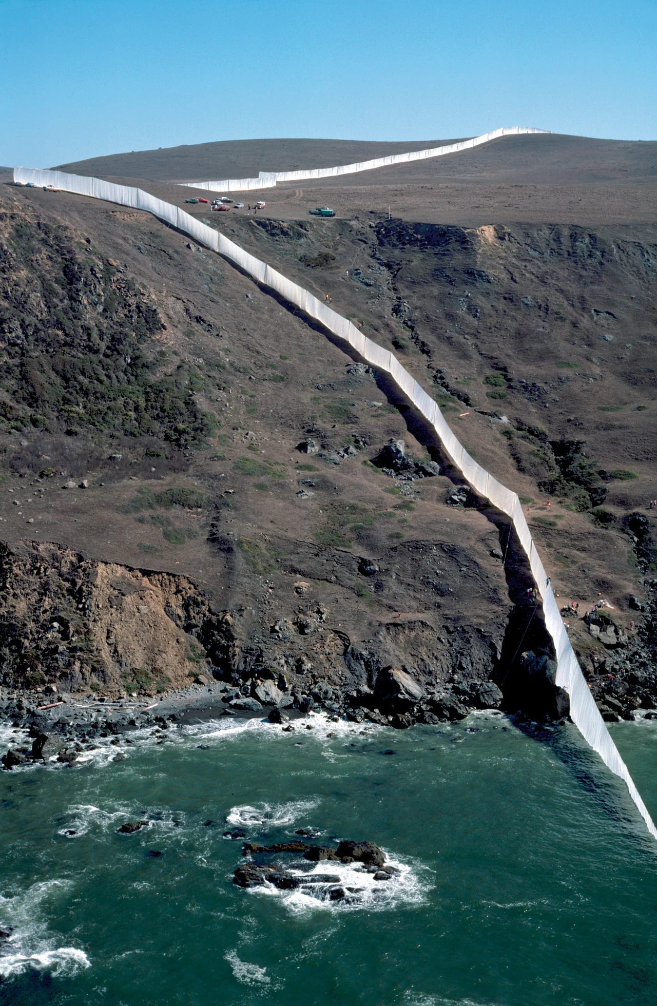 Seen from offshore, Running Fence crosses the California hills and runs down a rocky cliff into the surf.