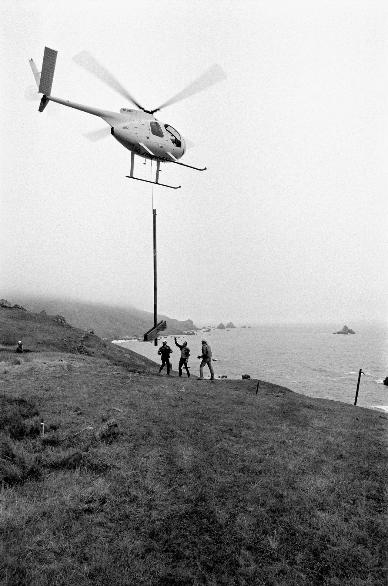 A helicopter lowers a steel pole toward workers on the California coast during the installation of Running Fence in 1976.